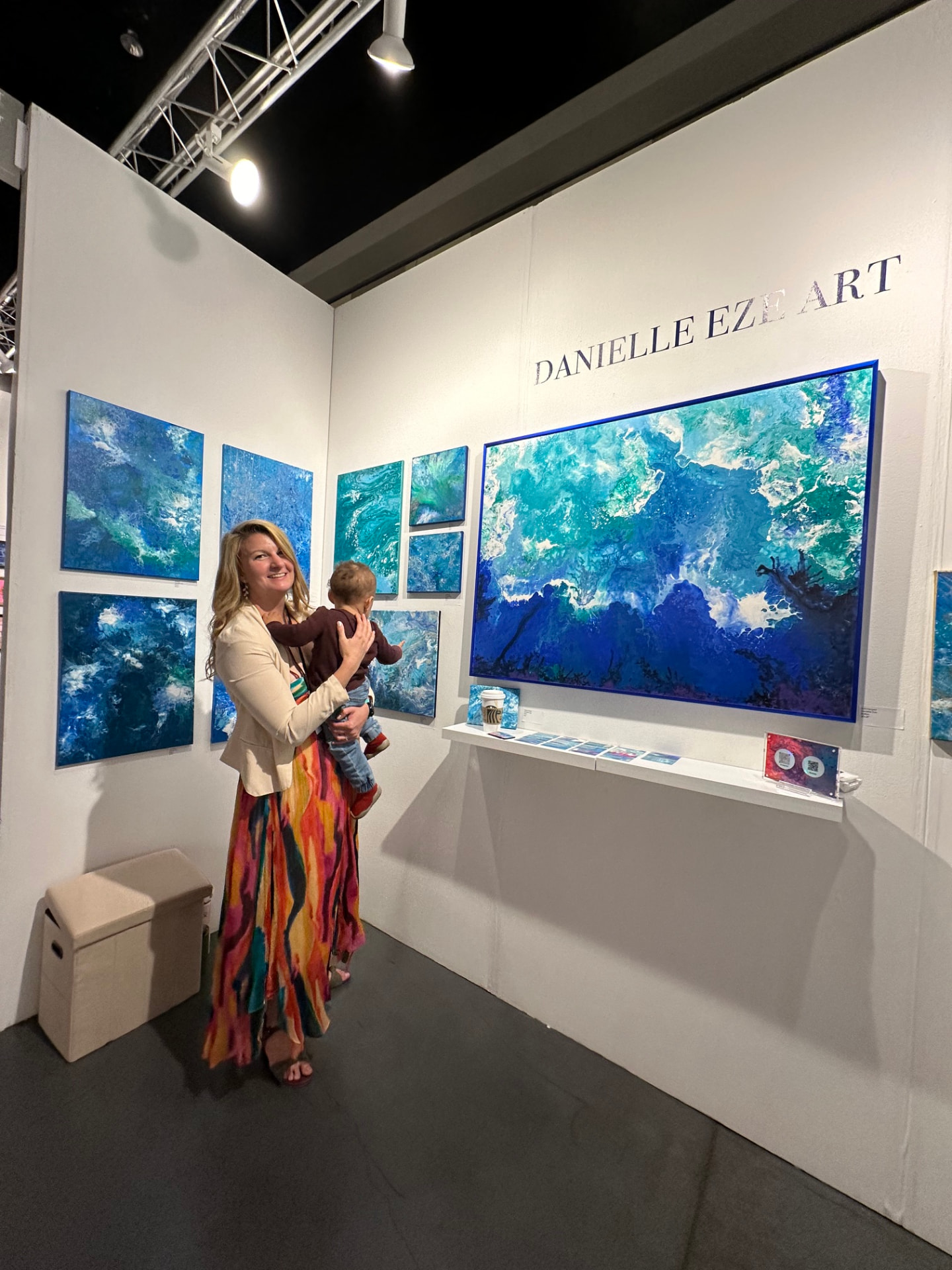 Danielle Eze with her child in front of a wall of blue ocean paintings at an art fair booth
